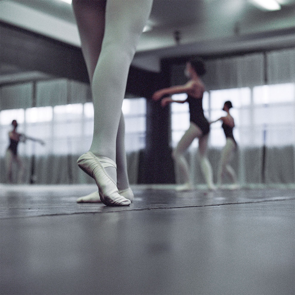 close-up of ballerina's legs in tights and ballet shoes on the dance floor with dancers blurred in the background.