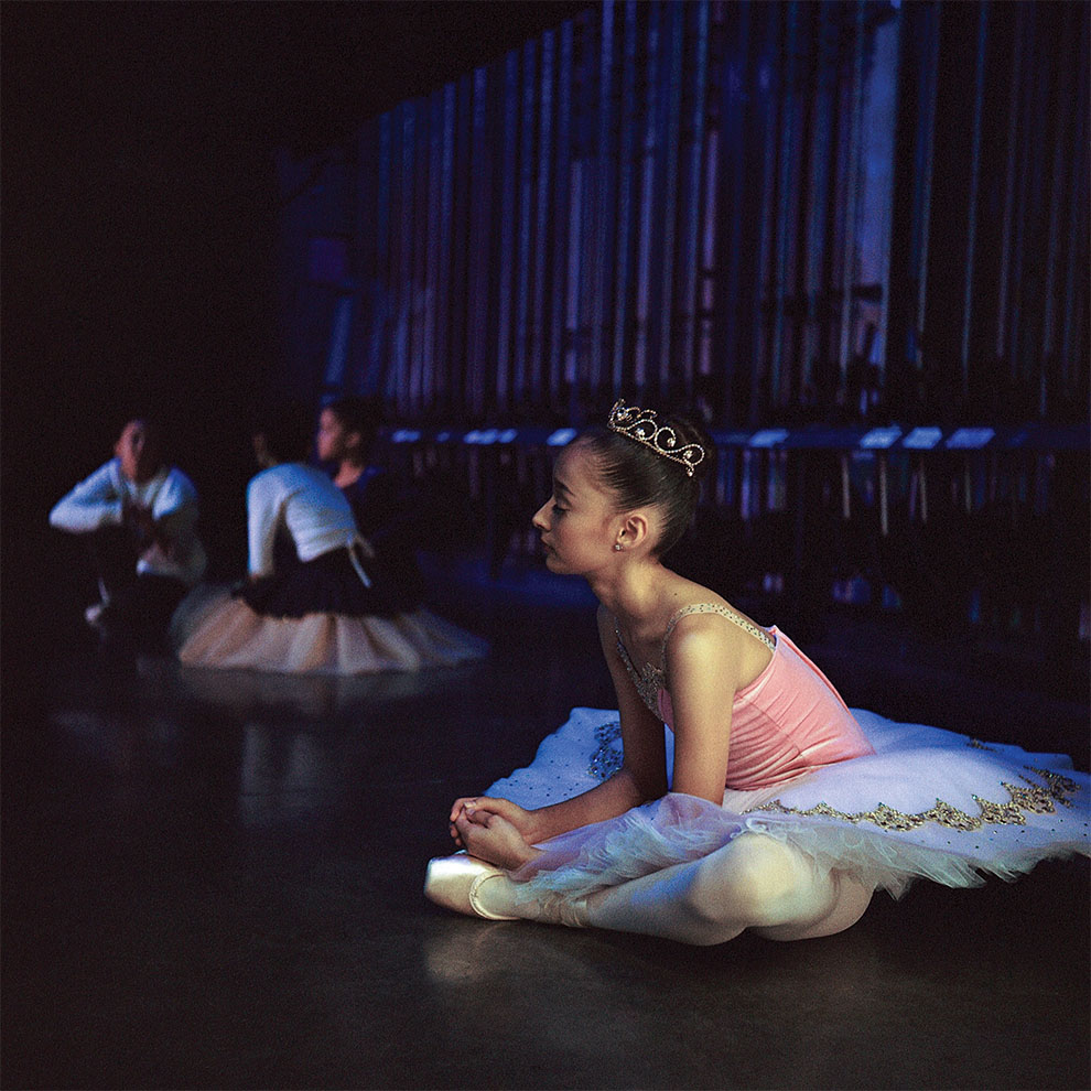 ballerina in tutu and tiara stretching sitting down backstage in the wings.