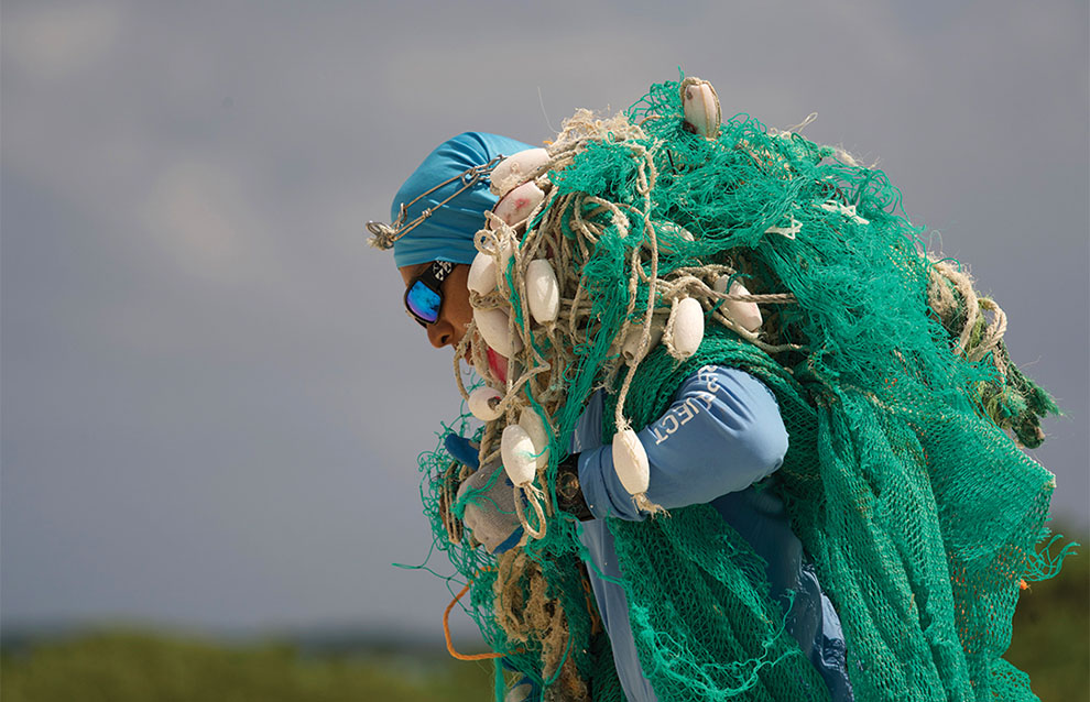 man in blue swimming cap and goggles pulls massive green net with rope across beach over his shoulder.