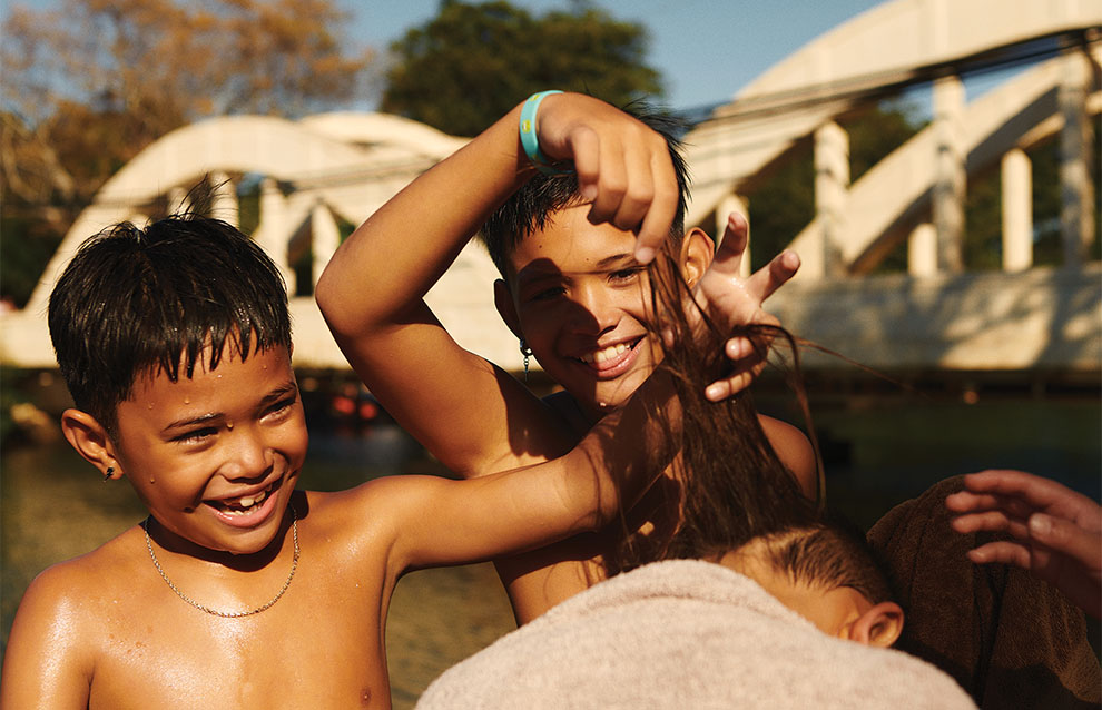 two young, tan boys have fun playing with another kid's hair by the bridge.