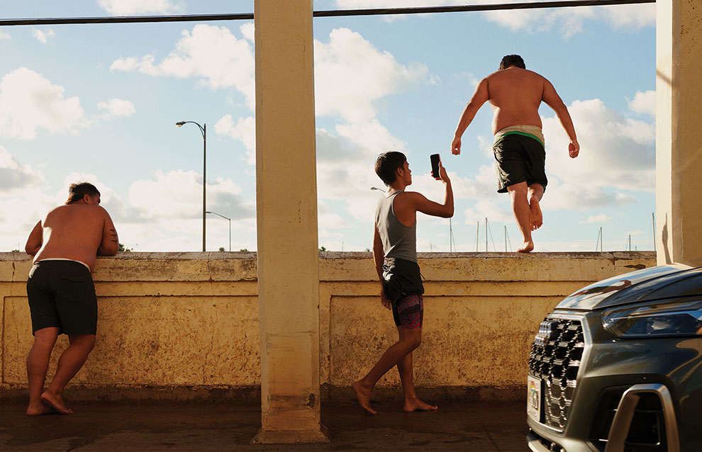 one person stands leaning against edge of bridge while another boy jumps off and his friend uses his phone to capture the moment.