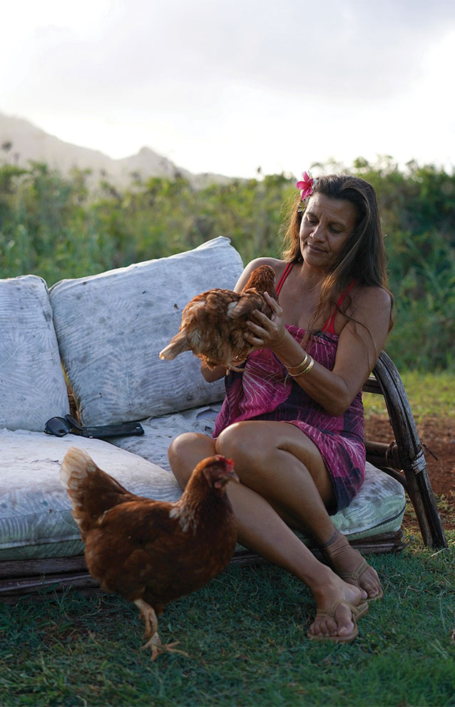 woman in pink dress sits on white cushion while holding up a chicken and another chicken stands by her legs.