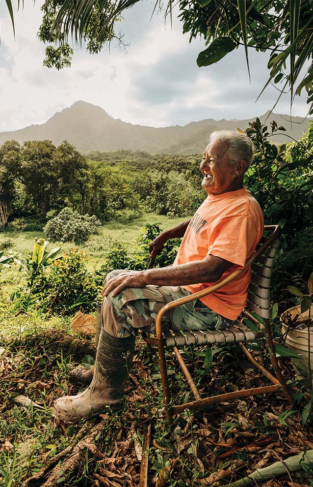 older man in bright orange t-shirt sits outside on a wooden chair with the Hawaiian mountains and lush landscape in the background.
